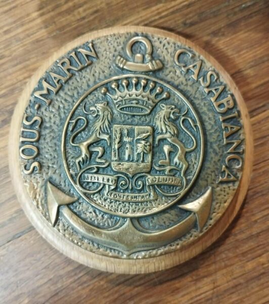 Round brass medallion with an ornate heraldic seal, featuring two lions supporting a shield and a crown, mounted on wood. The edge is engraved with a circular inscription.