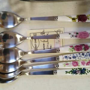 Six teaspoons with decorative floral porcelain handles laid in a row on a light table, showing pink, blue, and purple rose patterns as a set of flatware pieces.