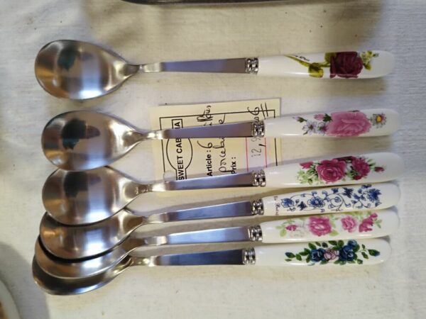 Six teaspoons with decorative floral porcelain handles laid in a row on a light table, showing pink, blue, and purple rose patterns as a set of flatware pieces.
